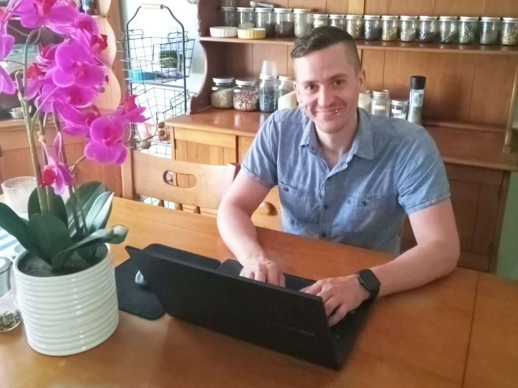 Dustin Sigsbee, the new assistant editor at Daily Nous, seated at a table in front of a laptop computer and a potted orchid.