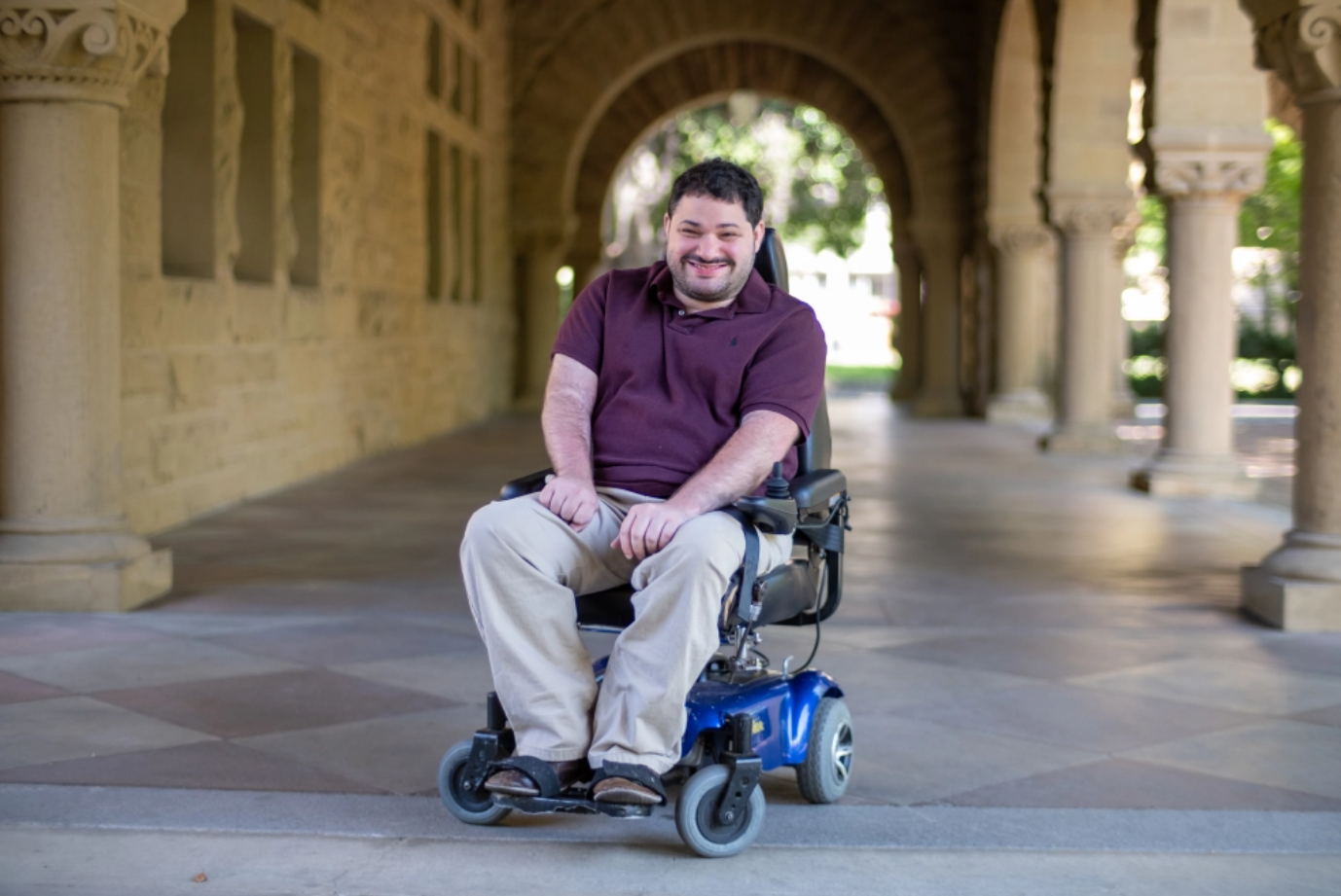 Photograph of Kevin Mintz amid columns and archways on the Stanford University campus, seated in a motorized wheelchair, smiling, and wearing a burgundy shirt and khaki pants.
